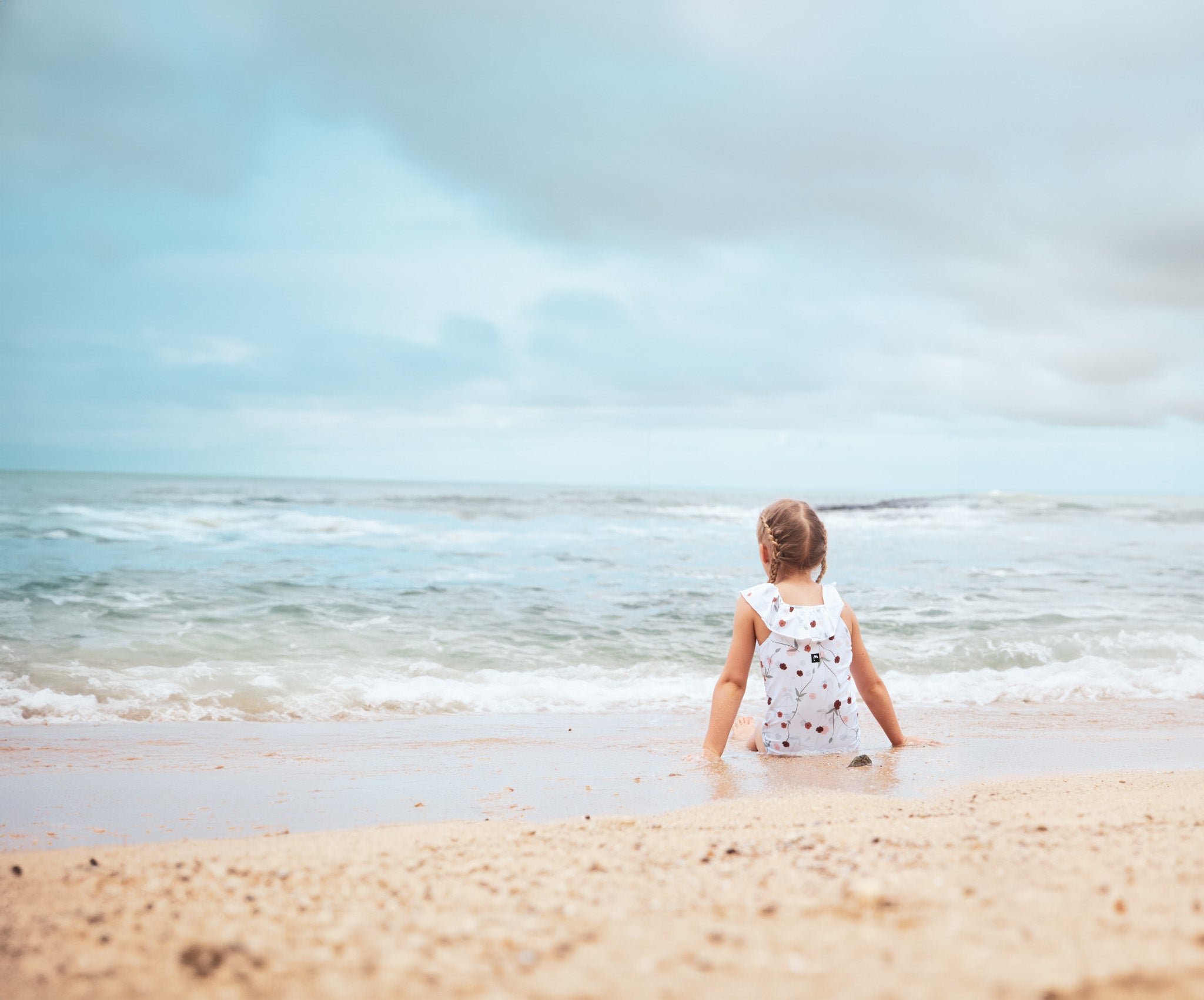 Toddler girl wearing KidORCA floral swimsuit at the beach, sitting near ocean waves